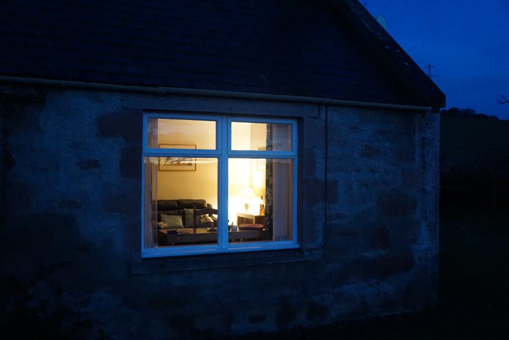 Ein warm erleuchtetes Fenster in einem alten Cottage bei blauer Stunde – ein Symbol für Stille, Rückzug und einen geschützten Ort der Einkehr.